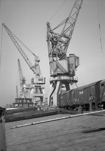 849773 Afbeelding van de overslag van goederen van trein naar schip in de haven te Rotterdam, met behulp van een havenkraan.